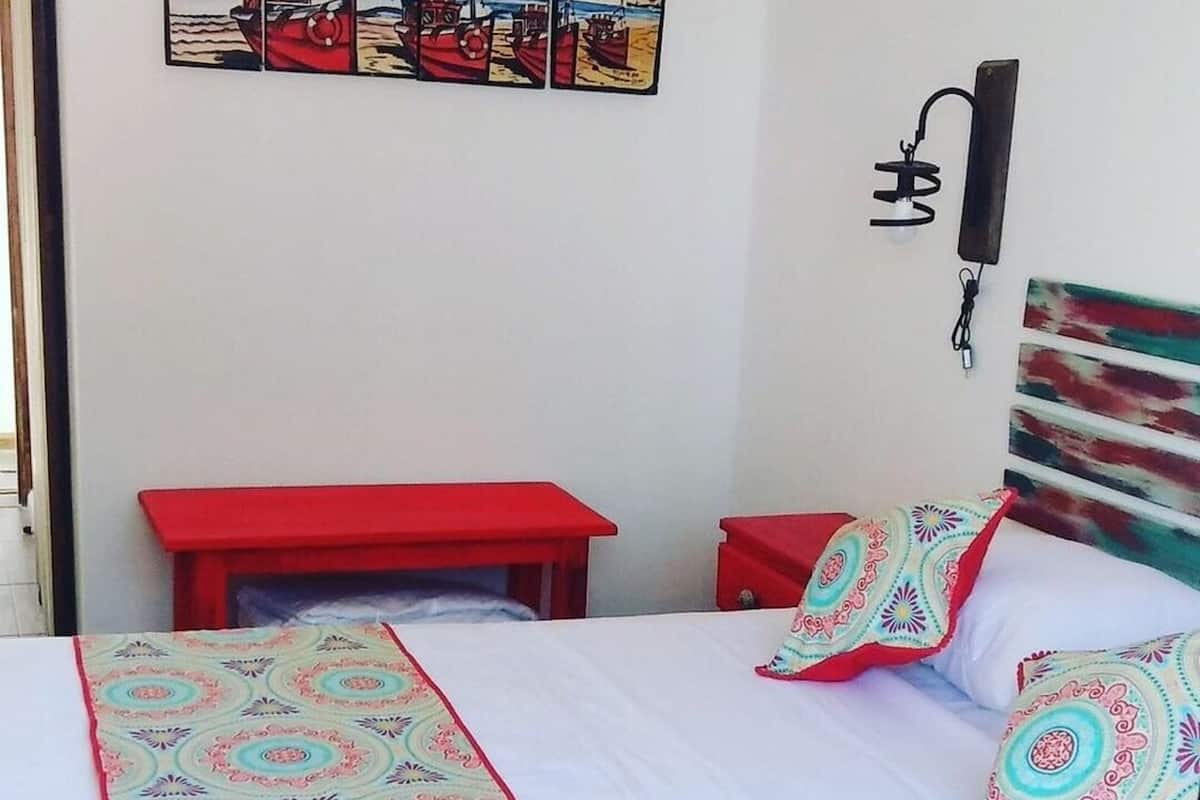 A bed with a patterned bedspread and pillows, a red bedside table, and a wall-mounted lamp.