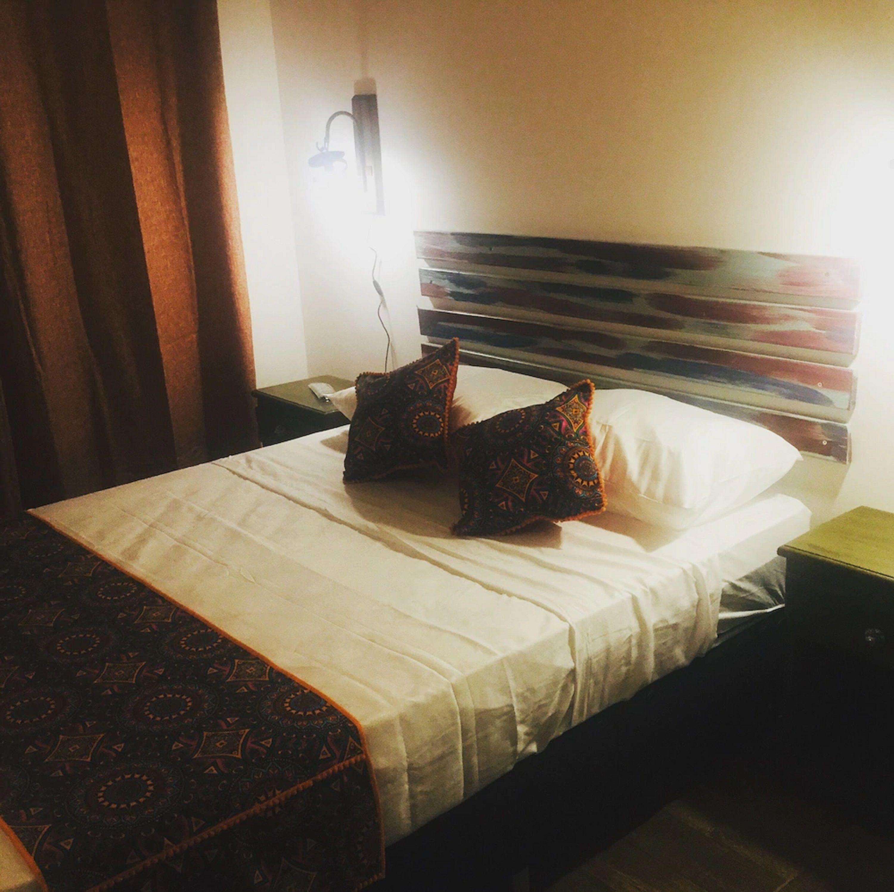Siisti hotellihuone, jossa on huolitellusti pedattu sänky, yöpöytä, seinään kiinnitetty lamppu ja kuviollinen torkkupeitto.