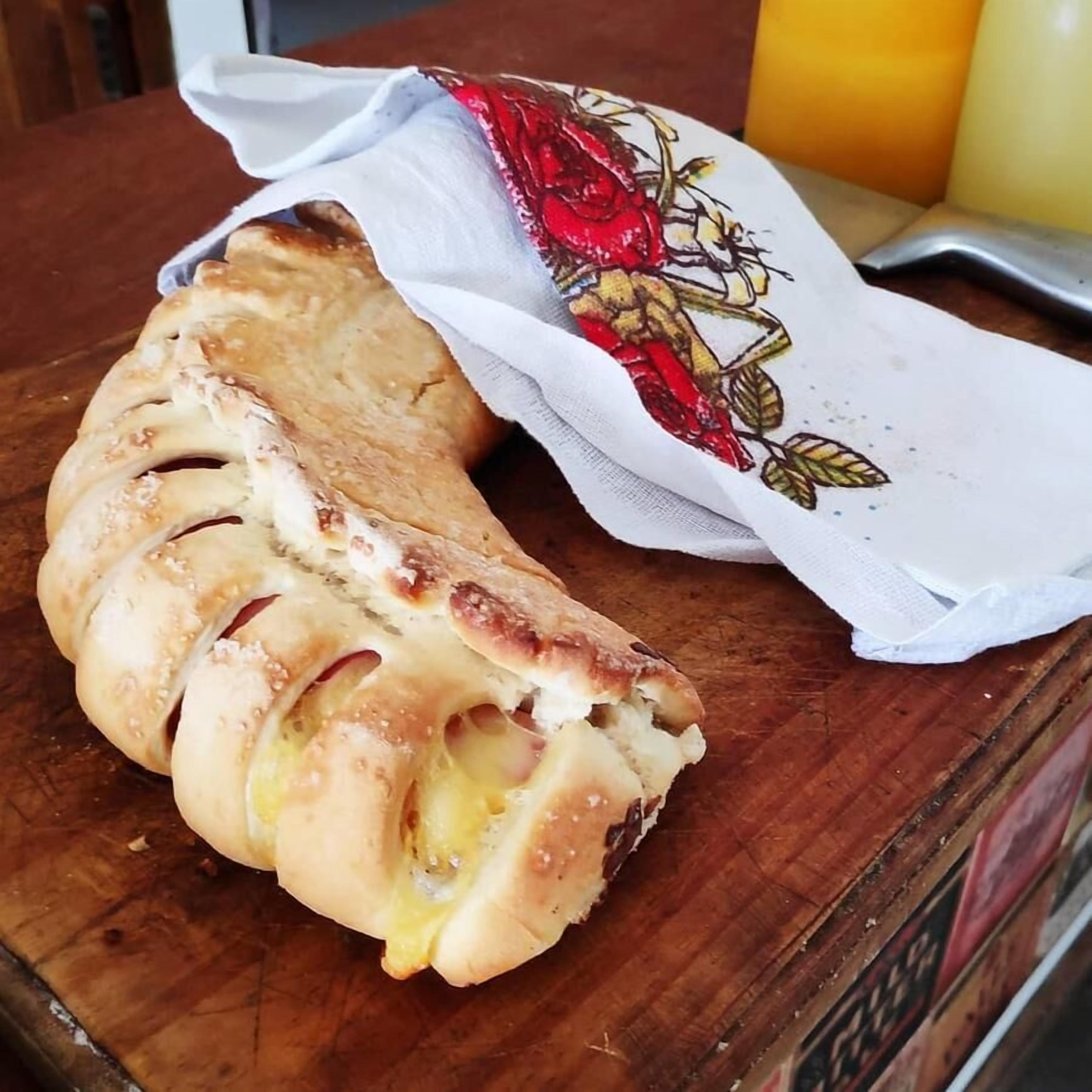 A loaf of bread with a crusty exterior and visible cheese filling, placed on a wooden surface next to a white cloth with a floral pattern.