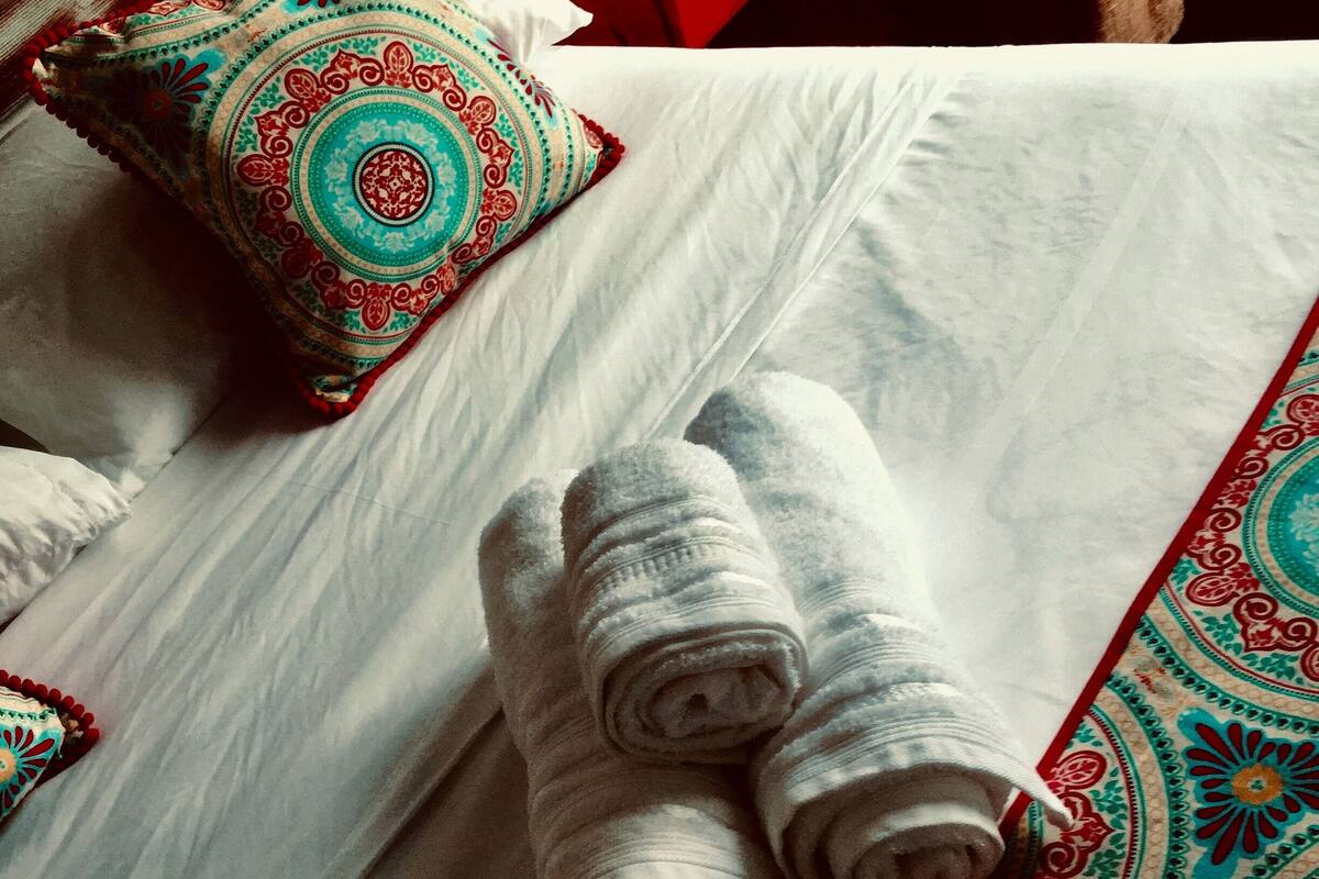 A neatly made bed with white linens, a decorative pillow, and a rolled-up towel on a patterned bedspread.