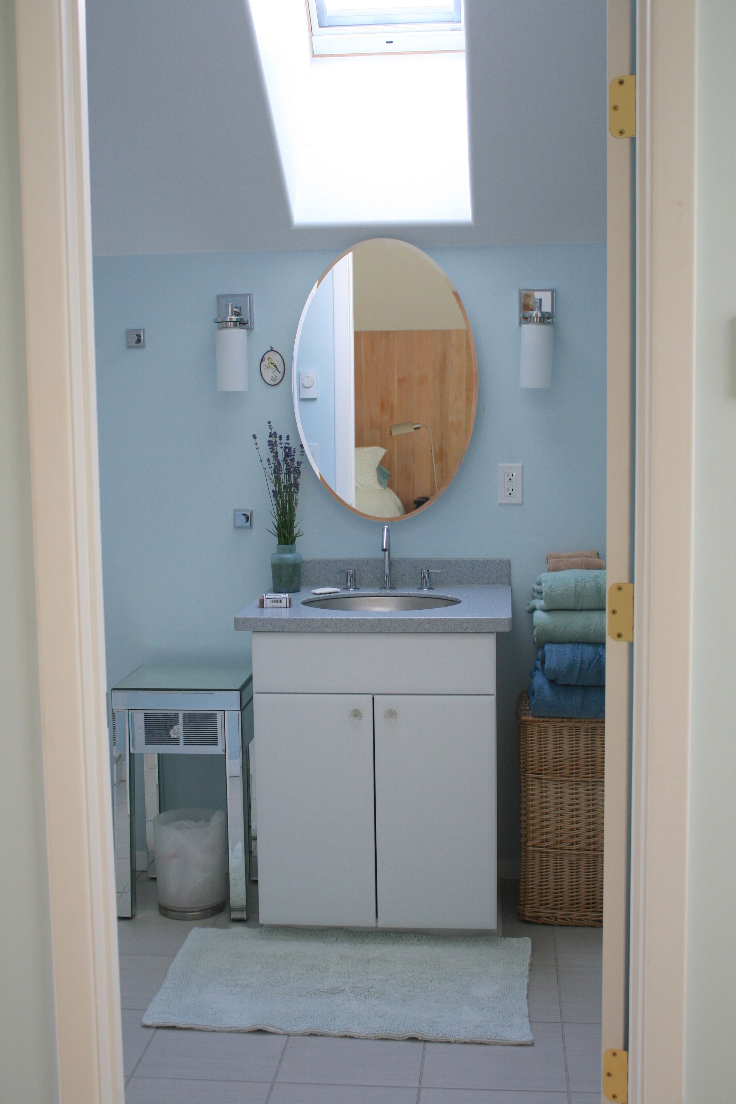 A bathroom with a skylight, a round mirror, a sink, and a small table with a plant.