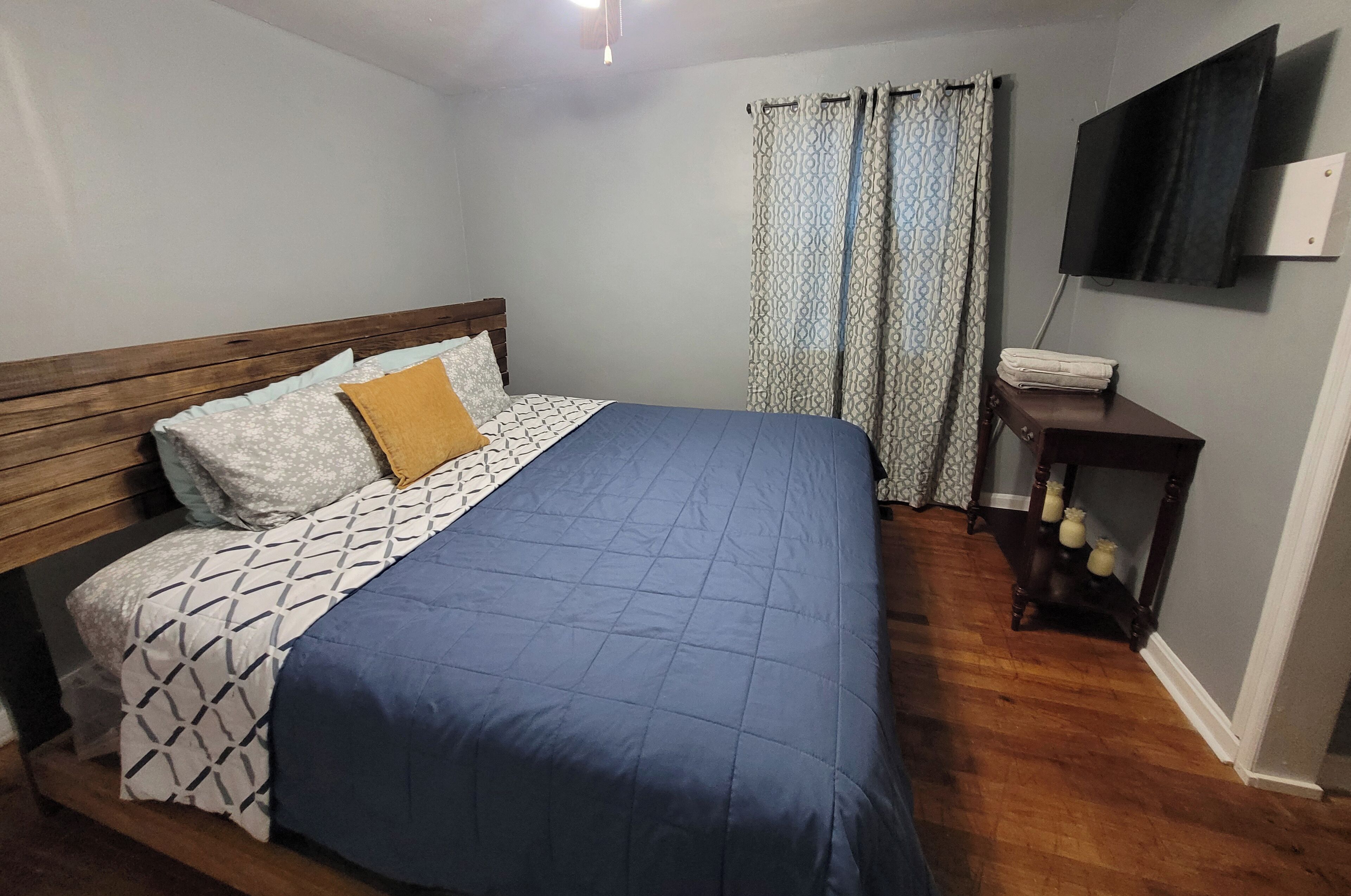 Ein Schlafzimmer mit Bett, Holz-Kopfteil, gemusterten Betttextilien, einer blauen Tagesdecke, einem Nachttisch mit Lampe und einem an der Wand montierten Fernseher.