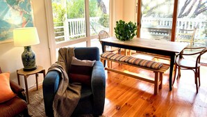 Een gezellige kamer met een houten eettafel, een comfortabele bank en een bankje. Er is een groot raam met uitzicht op een terras en bomen.