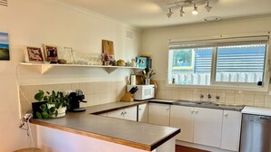 Een keuken met een centraal eiland, witte kastjes, een magnetron en een vaatwasser. Er is een raam met zonwering, een plant op het eiland en een klein krukje.