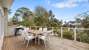 Une terrasse en bois, agrémentée d’une table et de chaises, entourée d’une balustrade blanche, et offrant une vue sur un quartier résidentiel avec des arbres et des maisons.