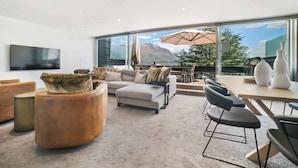 Ein modernes Wohnzimmer mit einer Couch, Hocker und Sesseln, einem Flachbildfernseher und einem großen Fenster mit Blick auf die Berge.