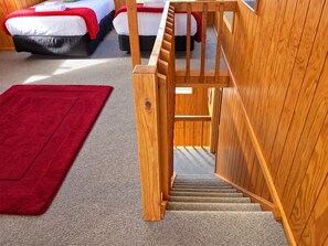 A hotel room with a wooden staircase leading to a balcony, a red carpet, and two beds with red and white bedding.