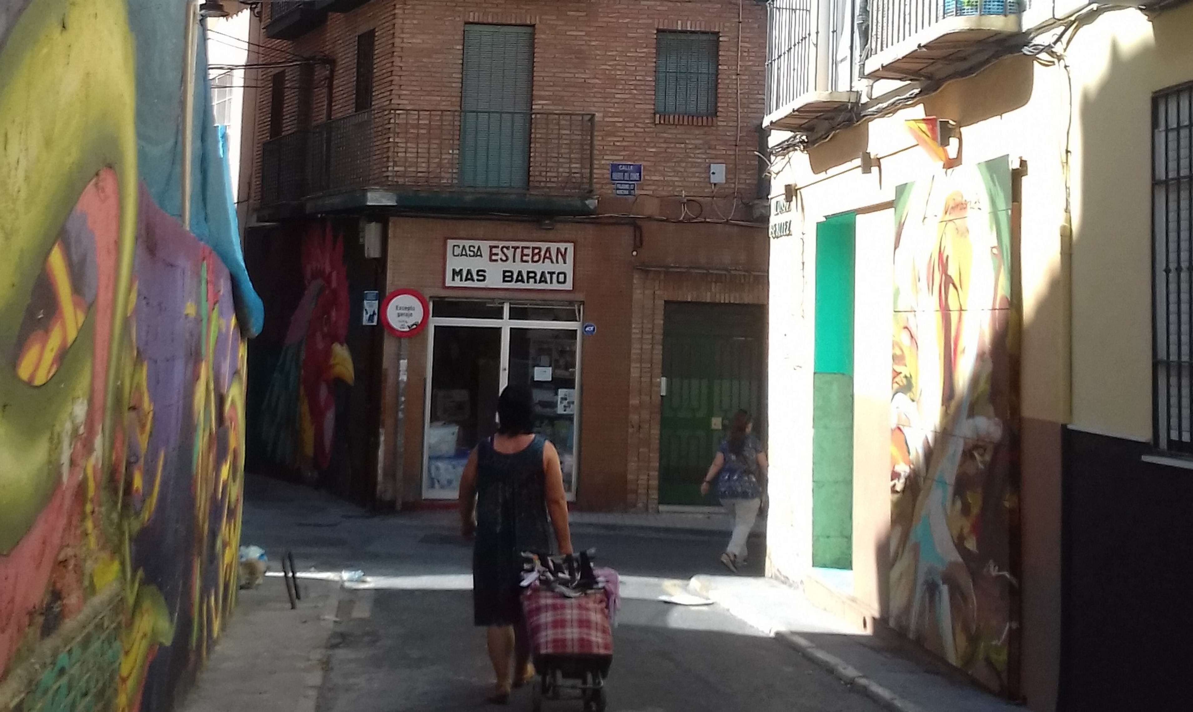 A narrow street with a building labeled 'CASA ESTEBAN MAS BARATO' and a mural on the left.