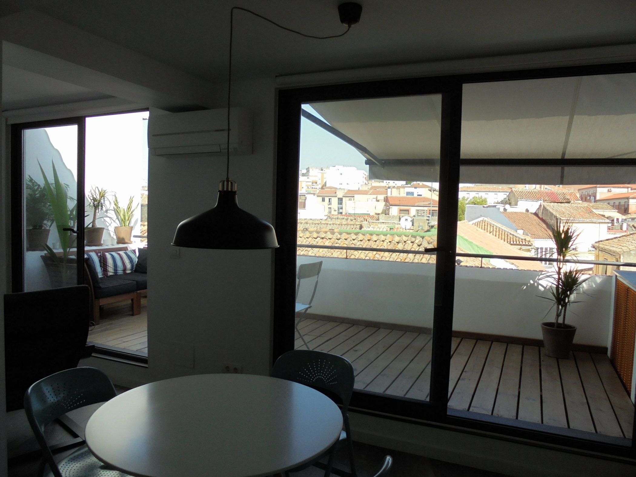A modern room with a round table and chairs, a hanging pendant light, and large windows offering a view of a cityscape.