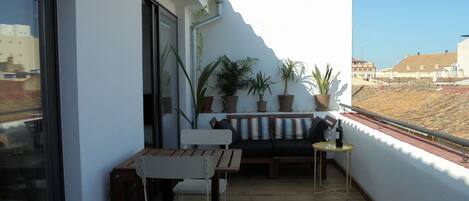 Ein Balkon mit Holztisch und -stühlen, Blumentöpfen und einem kleinen Tisch mit Flasche und Glas.