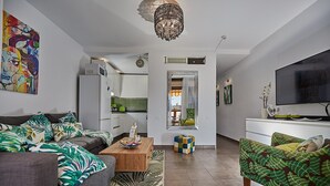 Un salon moderne avec un canapé, une table basse et un fauteuil coloré. On aperçoit une télévision et une cuisine au fond de la pièce.