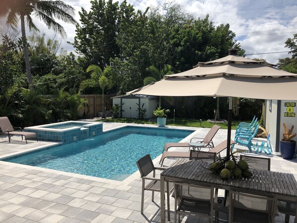 A rectangular swimming pool with tiled steps, surrounded by lounge chairs and a table with a canopy.