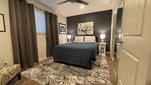 A bedroom with a bed, a ceiling fan, a nightstand with a lamp, a painting on the wall, and a door.