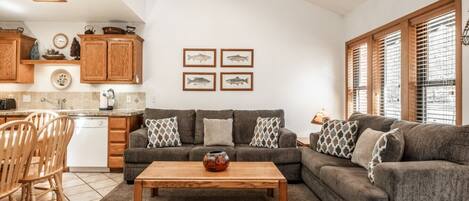 A cozy living room with a gray sectional sofa, wooden coffee table, and a kitchen area with wooden cabinets and a tiled backsplash.