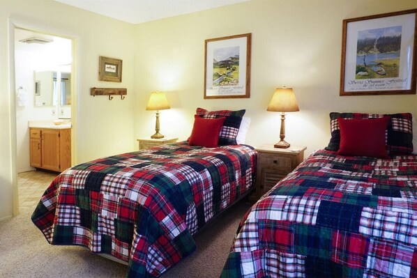 A room with two beds, each with a plaid bedspread, two bedside lamps, a wooden nightstand, a framed picture on the wall, and a bathroom visible through an open door.