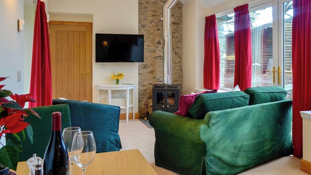 A cozy living room with a stone wall, a fireplace, a flat-screen TV, and a large window with red curtains. - The Old Inverchroskie Kennels (Enochdhu, near Pitlochry)