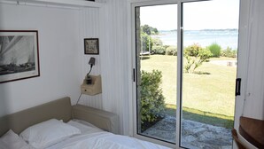 Ein Schlafzimmer mit Bett, Nachttisch, einem gerahmten Bild und Blick auf einen Garten und Wasser durch eine Schiebetür.