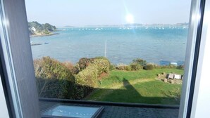 Blick aus einem Fenster auf ein Gewässer mit Booten, eine Wiese und eine entfernte Küste.