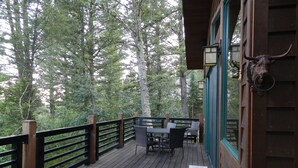 A wooden deck with a table and chairs, surrounded by a black railing, overlooking a forested area.
