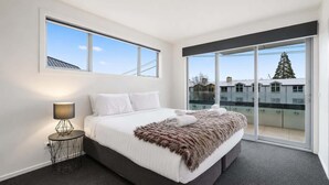A modern bedroom with a large bed, a nightstand, a lamp, and a view of buildings through large windows.