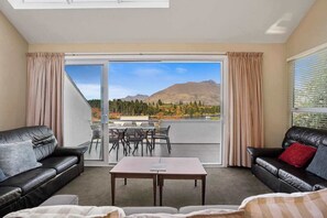 Quarto com porta de vidro deslizante para um pátio externo com vista para as montanhas. No interior, dois sofás de couro preto, uma mesinha e uma janela com persianas.