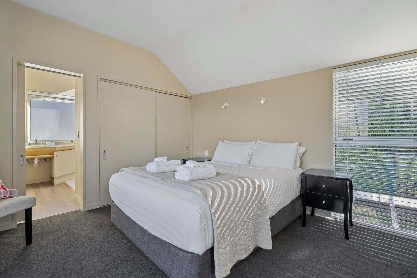 A hotel room with a bed, bedside table, and a window with blinds.