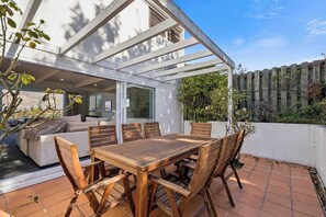 Eine Terrasse mit Holztisch und -stühlen, einer weißen Wand und Blick auf ein Gebäude.