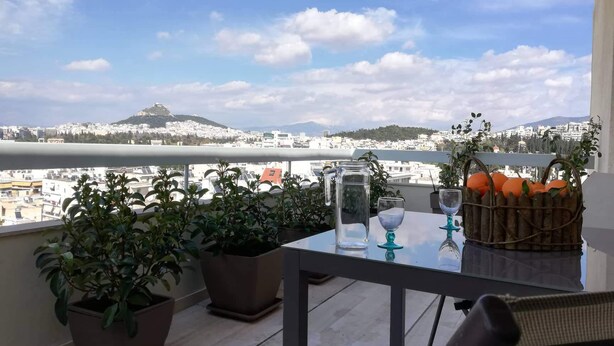 Uma varanda com mesa, plantas em vasos, um jarro e uma cesta de laranjas.