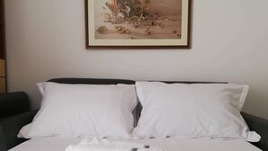 Cama bem arrumada com lençóis brancos e duas toalhas enroladas, uma obra de arte emoldurada acima da cama e cabeceira escura.