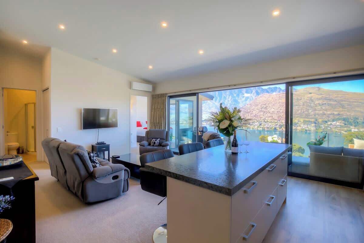 Ein modernes Wohnzimmer mit einer großen Kochinsel, bequemen Sitzgelegenheiten, einem Flachbildfernseher und einem weiten Ausblick durch große Fenster.
