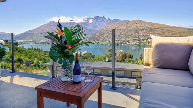 A terrace with a wooden table, a bottle of wine, and a glass, overlooking a lake and mountains.