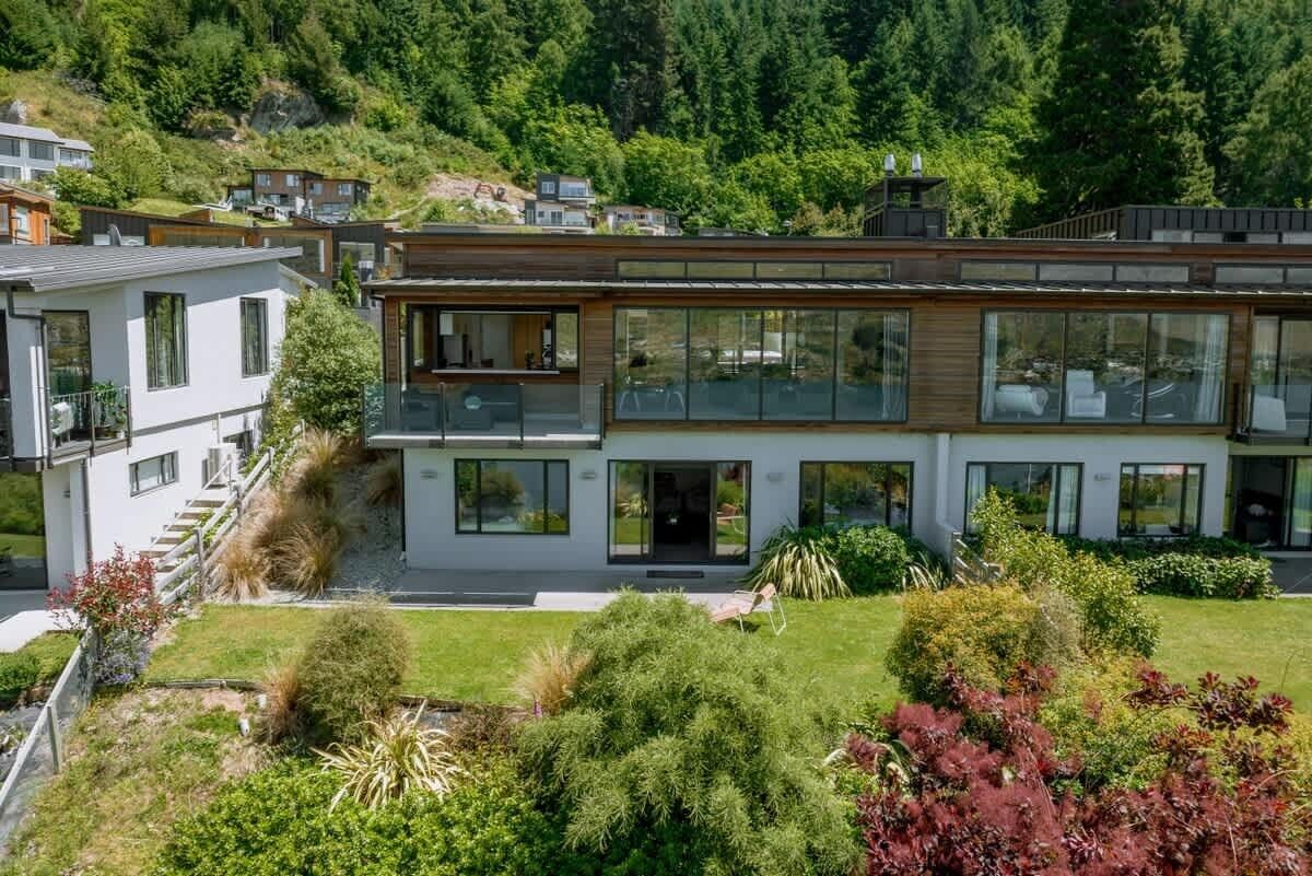 A modern two-story house with large windows and a balcony, surrounded by lush greenery and a hilly landscape.