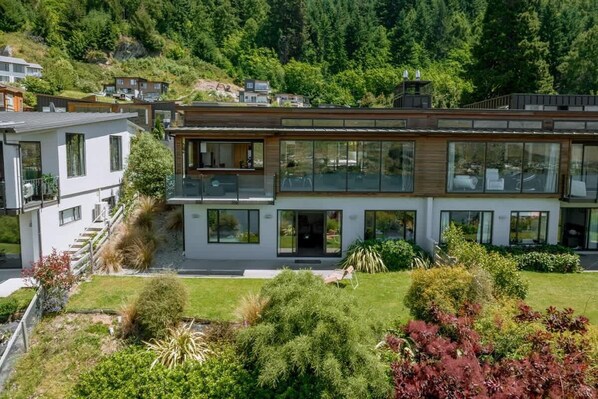 A modern two-story house with large windows and a balcony, surrounded by lush greenery and a hilly landscape.