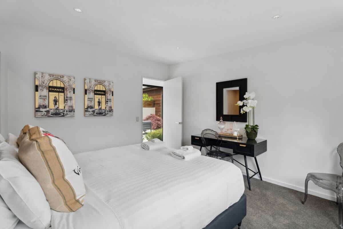 A modern hotel room with a large bed, a desk, and a chair. There are two framed pictures on the wall, a mirror above the desk, and a window with a view of greenery.