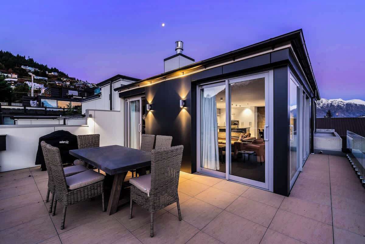 A modern outdoor dining area with a table and chairs, a barbecue grill, and a sliding glass door leading to a kitchen.
