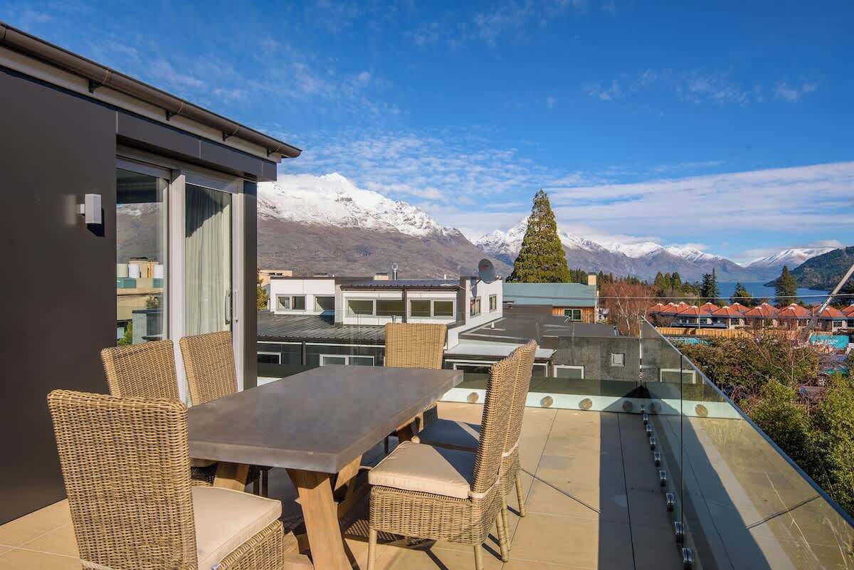 Eine Terrasse mit Tisch und Stühlen, mit Blick auf eine gebirgige Landschaft mit schneebedeckten Gipfeln.