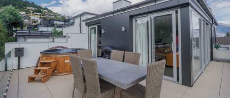 Een modern terras buiten met een eettafel, rieten stoelen en een jacuzzi.