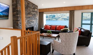 A modern living room with a stone fireplace, a black leather sofa with red cushions, a patterned armchair, and a large window overlooking a lake.