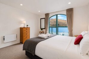 A hotel room with a bed, a dresser, a lamp, a view of the outdoors, and a window with curtains.