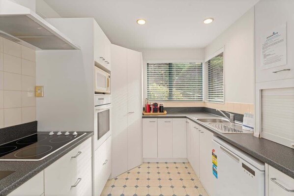 A compact kitchen with white cabinets, a black countertop, a built-in oven, and a window with blinds. - Remarkables Retreat 10 Lake Views & Balcony (Queenstown)