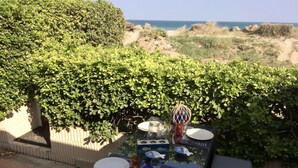 An outdoor dining table with plates, glasses, and cutlery set against a backdrop of greenery and a view of the sea.