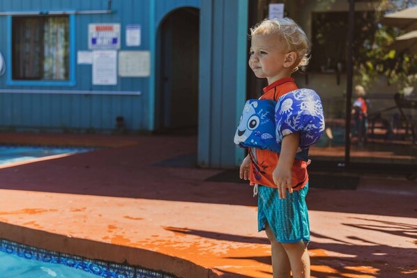 Niño de pie junto a una piscina, con un edificio azul al fondo.