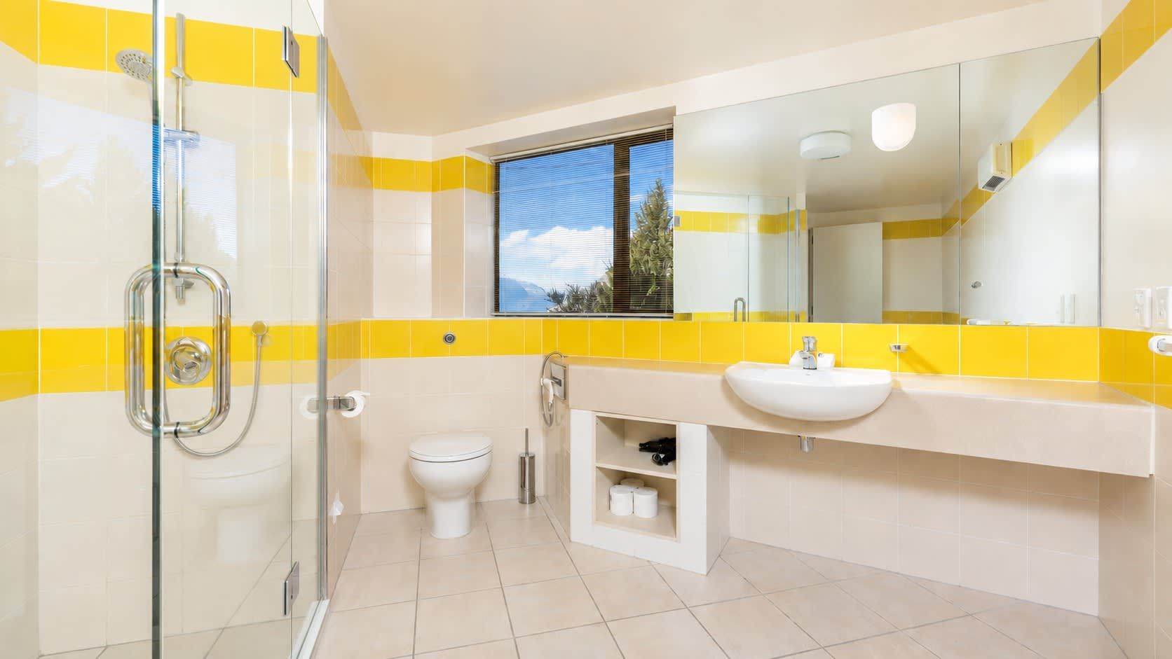 A bathroom with a glass shower enclosure, a toilet, a sink with a mirror above it, and a window providing a view of the outdoors.