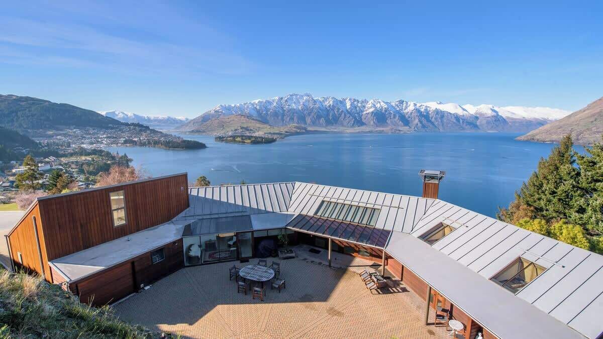 Un bâtiment moderne doté d’un toit en pente, d’un patio aménagé avec du mobilier d’extérieur, et offrant une vue sur un lac et des montagnes en arrière-plan.