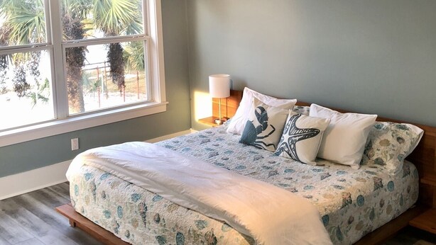 A bedroom with a bed, a nightstand, a lamp, and a window with a view of palm trees. - A Santa Rosa Sound Waterfront home on 2 acres with a private beach and 200’ dock (Mary Esther)