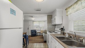 A compact kitchen with white cabinets, a double sink, and a microwave. There is a small dining area with a chair and table, and a window with blinds.