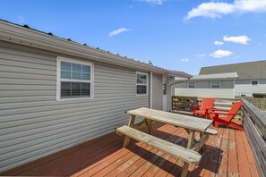 Una terraza de madera con mesas de picnic y sillas Adirondack rojas, junto a una casa con revestimiento blanco y techo a dos aguas.