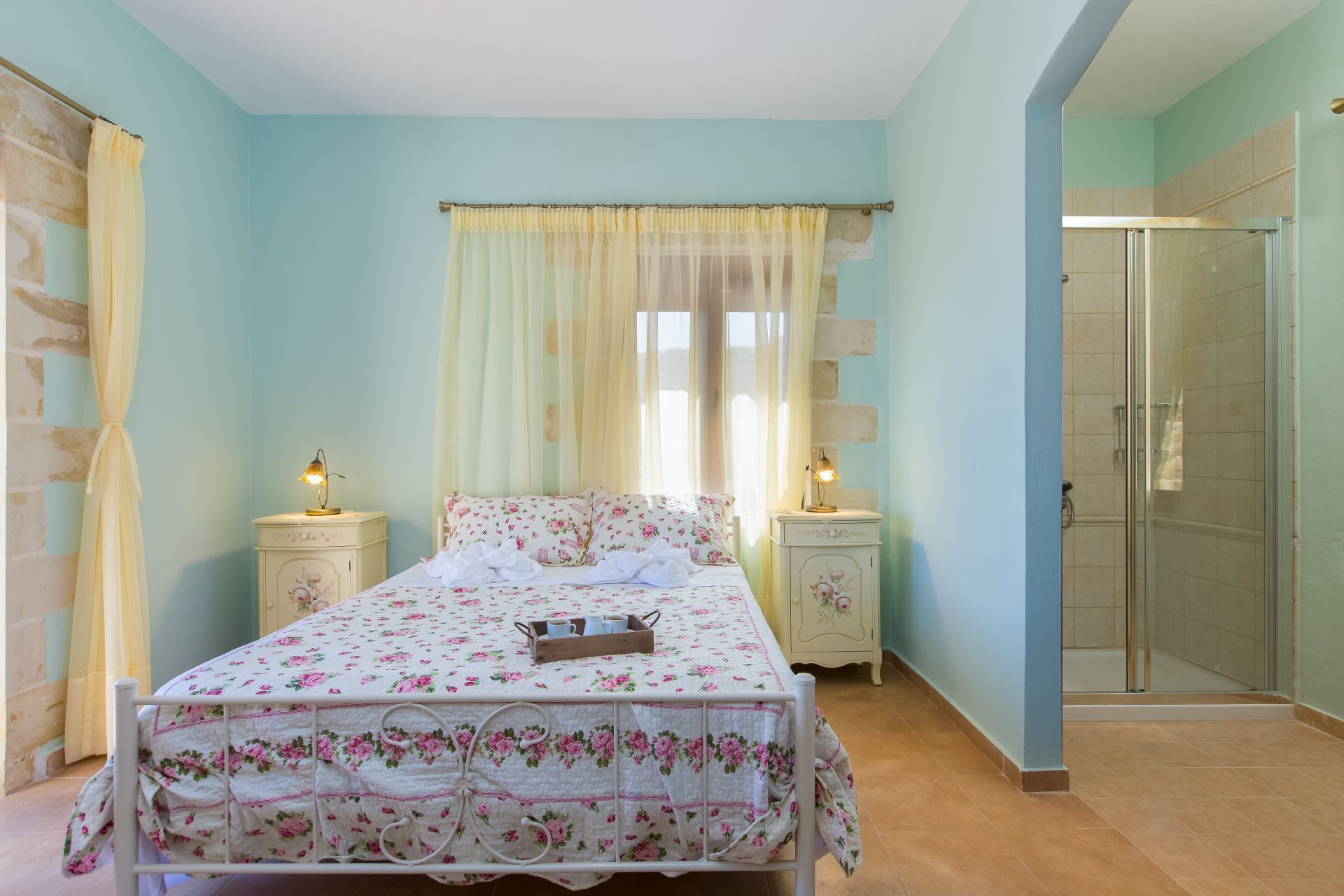 Un dormitorio con cama, dos mesitas de noche, ventana con cortinas translúcidas, zona de ducha y puerta que da a otra habitación.