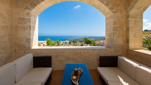 A stone-walled room with a view of the sea. There is a table with a blue tablecloth and two cushioned benches. - Faethon, eco stone villa with private pool and sea view (Rethymno)