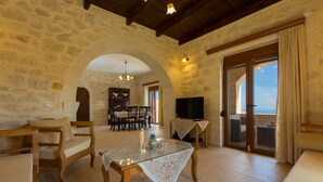 A cozy room with stone walls, a dining area with chairs and a table, a TV, and a balcony with curtains. - Faethon, eco stone villa with private pool and sea view (Rethymno)